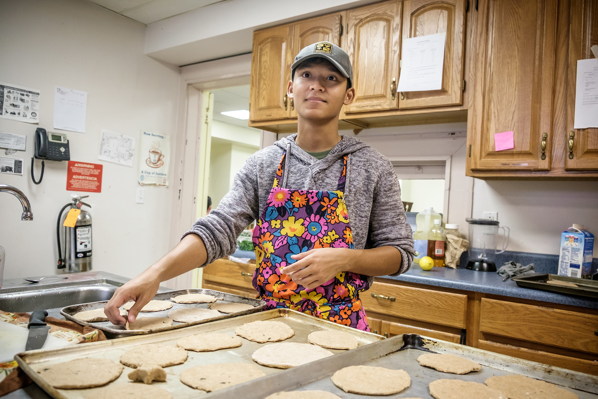 man with a floral apron on in the kitchen putting flattened cookies on a cookie tray