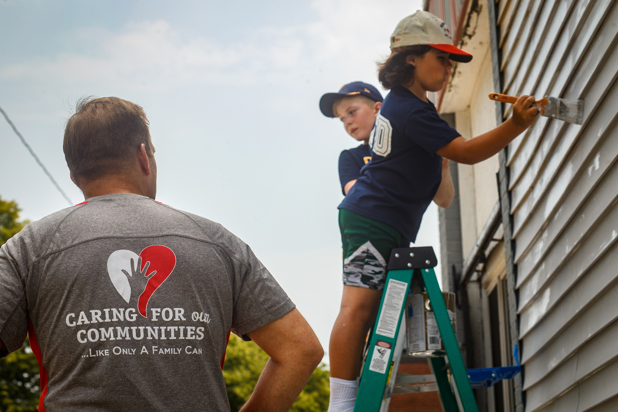 two student volunteers on a ladder painting a house while a man supervises